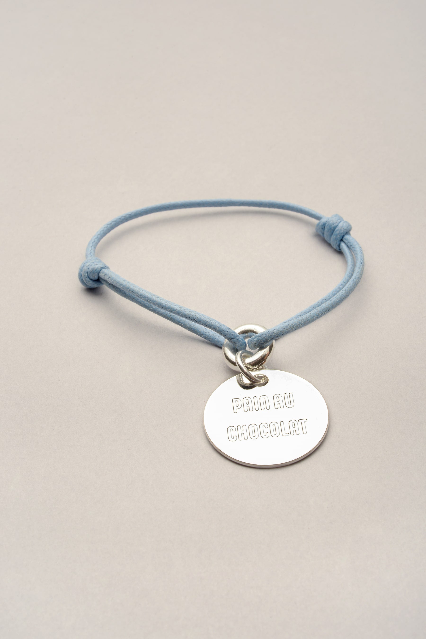Ce bracelet cordon est composé d'une médaille sur laquelle est gravée "pain au chocolat". Bijou fantaisie argenté réglable grâce à deux noeuds coulissants. Fabriqué en France. 