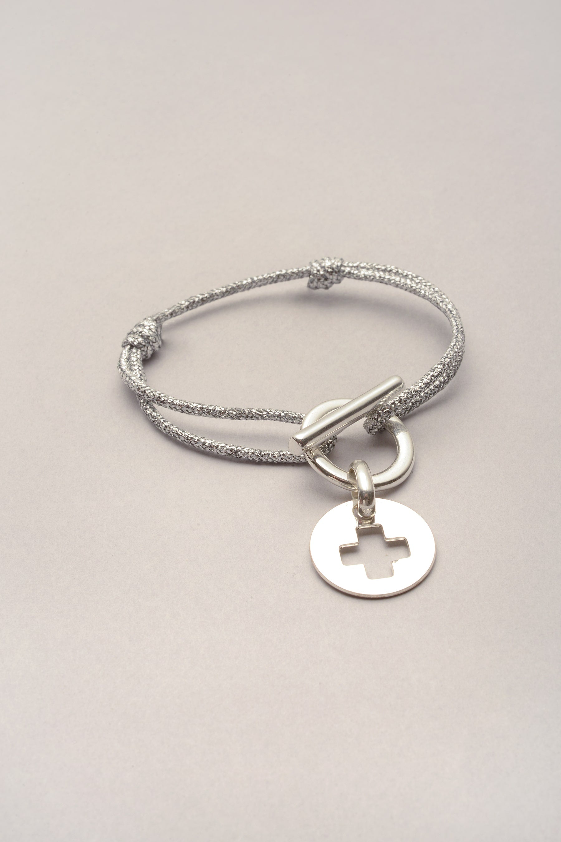 Ce bracelet est composé d'un cordon de coton coloré, d'un fermoir en T et d'une médaille avec une croix évidée. Ce bijou s'attache et se ferme grâce à son fermoir, ce qui rend pratique l'utilisation ! 