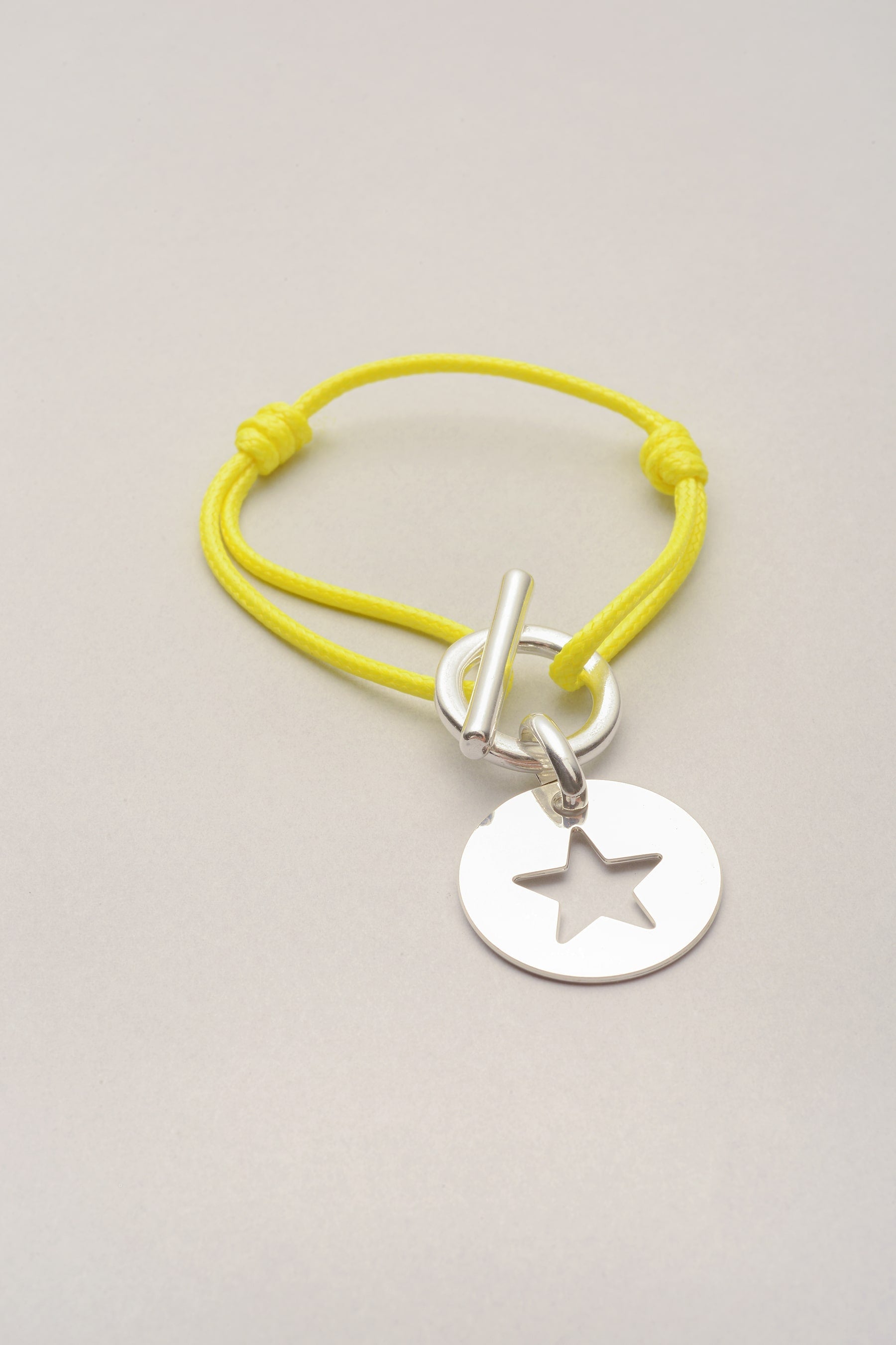 Ce bracelet est composé d'un cordon de coton coloré et d'un fermoir en T. Sur le fermoir, une médaille avec une étoile évidée est présente. Ajustable, ce bracelet fantaisie argenté se règle grâce à deux noeuds coulissants. 