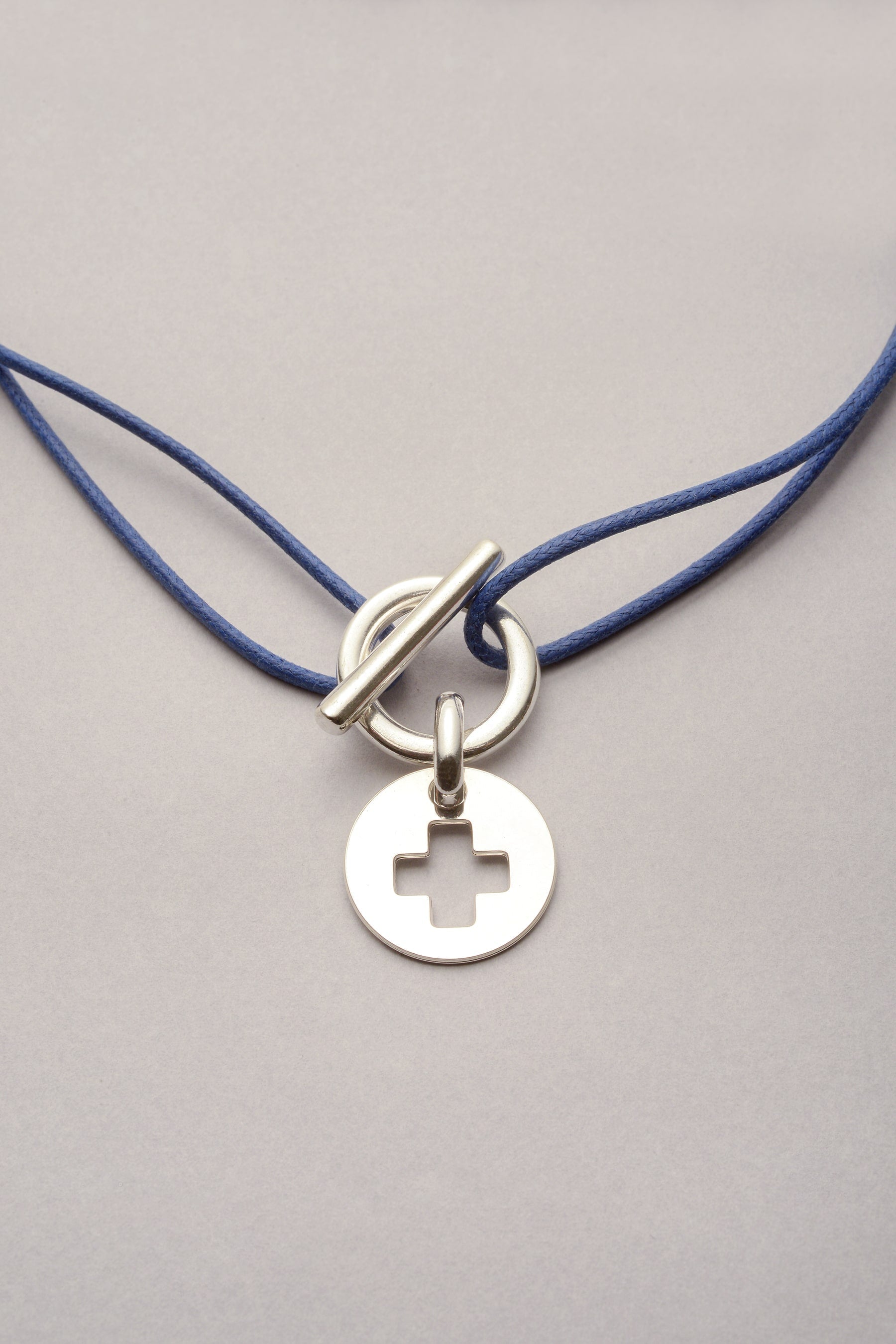 ce collier est composé d'un cordon de coton coloré et d'une attache. L'attache est un fermoir en T, qui permet l'ouverture et fermeture facile du bijou. Sur le fermoir en T, il y a une médaille avec une croix en forme de + évidée. 