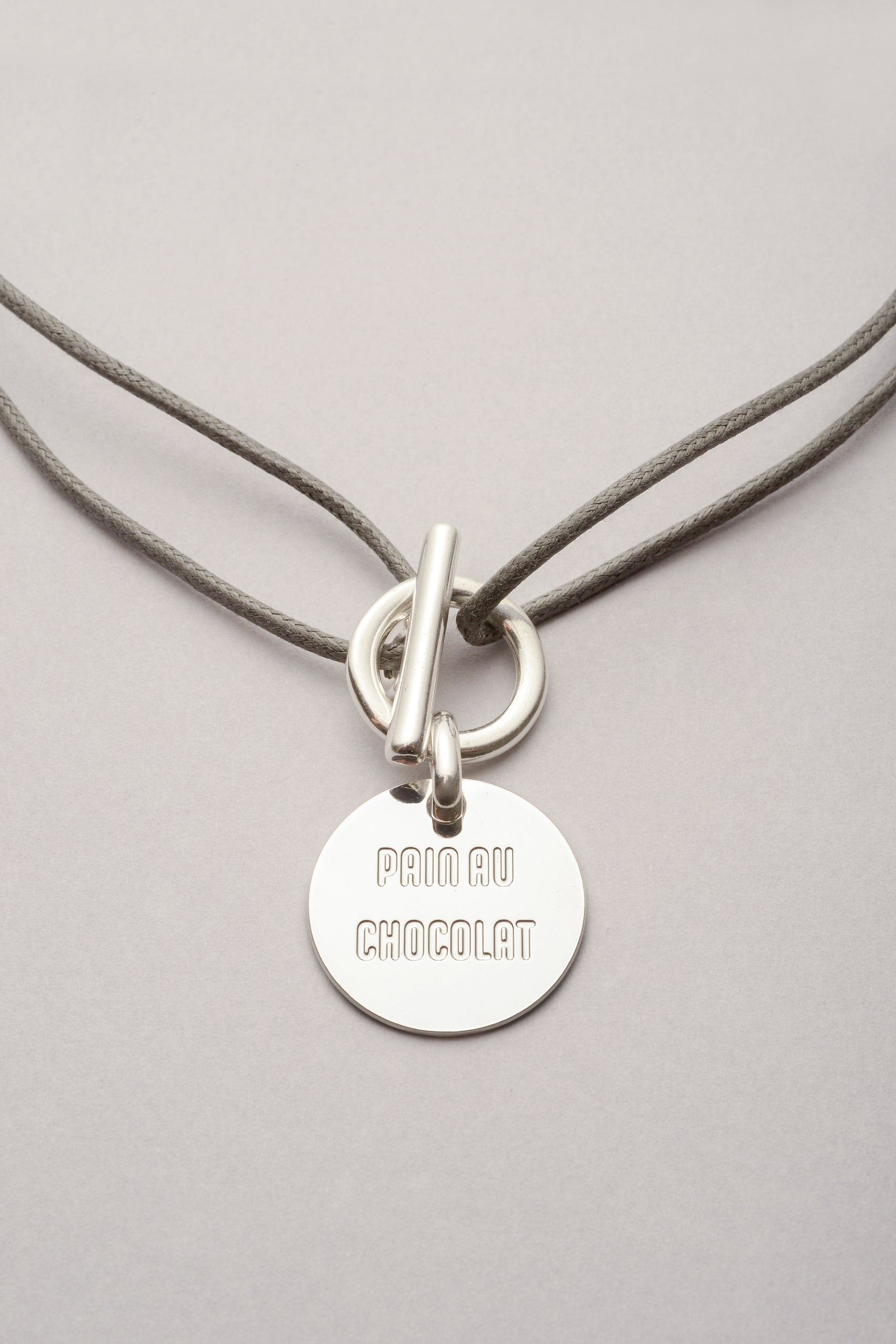 Ce collier est composé d'un cordon et d'une attache sur laquelle est accrochée une médaille. Sur la médaille, on lit "pain au chocolat". L'attache est un fermoir en T, ce qui rend facile l'utilisation du bijou. Le collier attache médaille “pain au chocolat” est une création Musardise pleine de caractère : argenté, réglable et 100 % made in Toulouse.