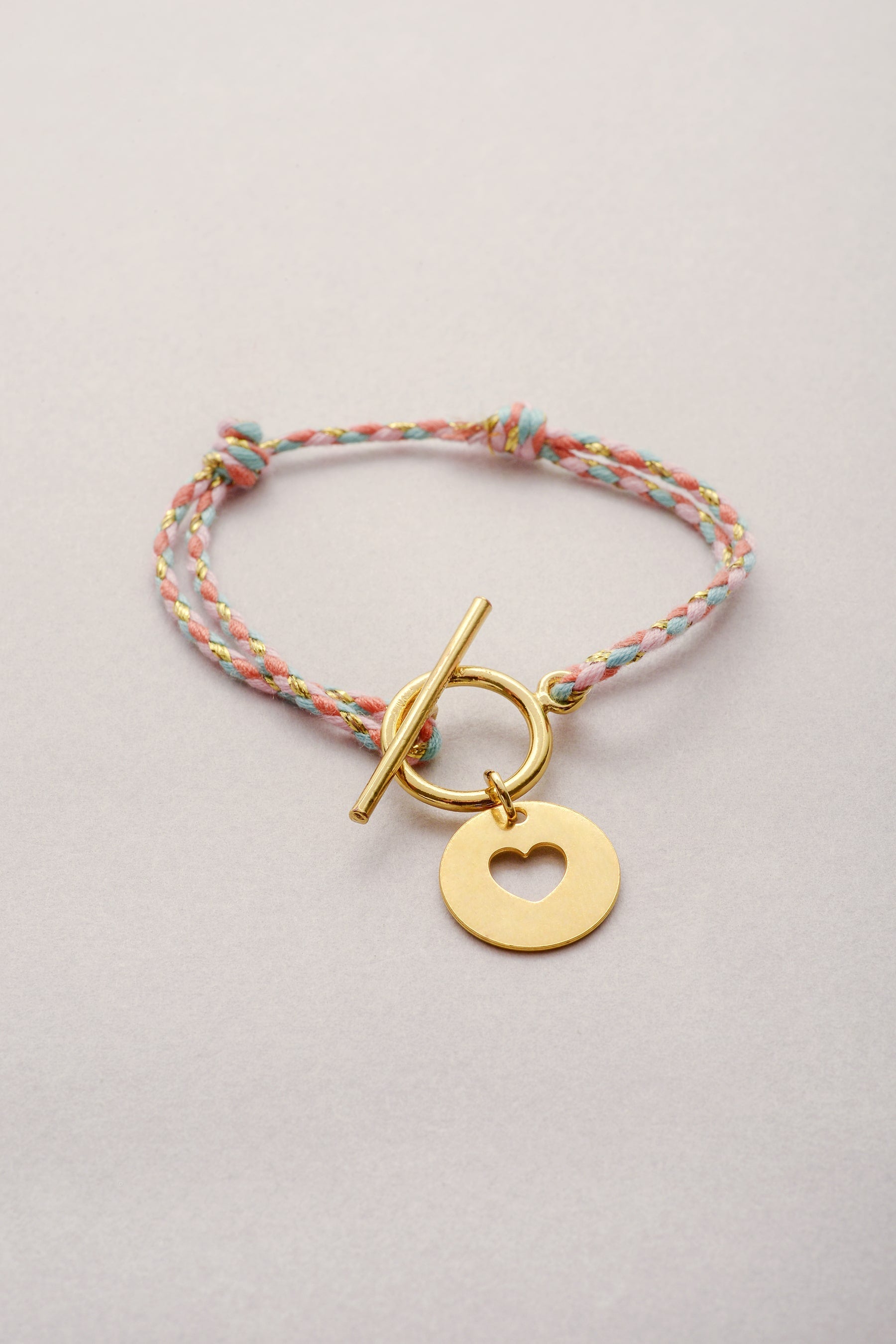 Bracelet en cordon tressé coloré avec sa pampille dorée en forme de coeur ajouré et son fermoir en T.