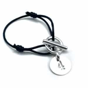 Bracelet argenté avec une attache et un fermoir en T. Il comporte également une pampille chiffre et un cordon en coton noir